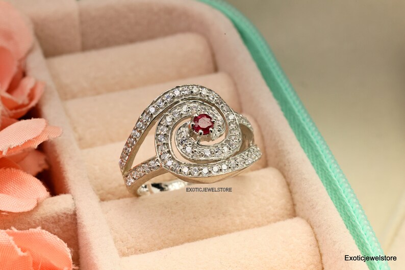 May include: A silver ring with a red gemstone in the center, surrounded by smaller white gemstones. The ring is set in a swirl design.