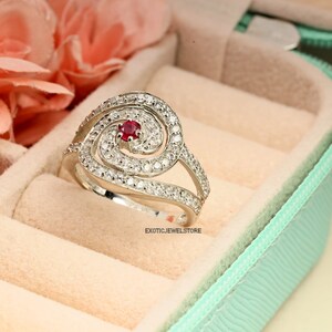 May include: A silver ring with a spiral design and a red gemstone in the center. The ring is set with small white gemstones.