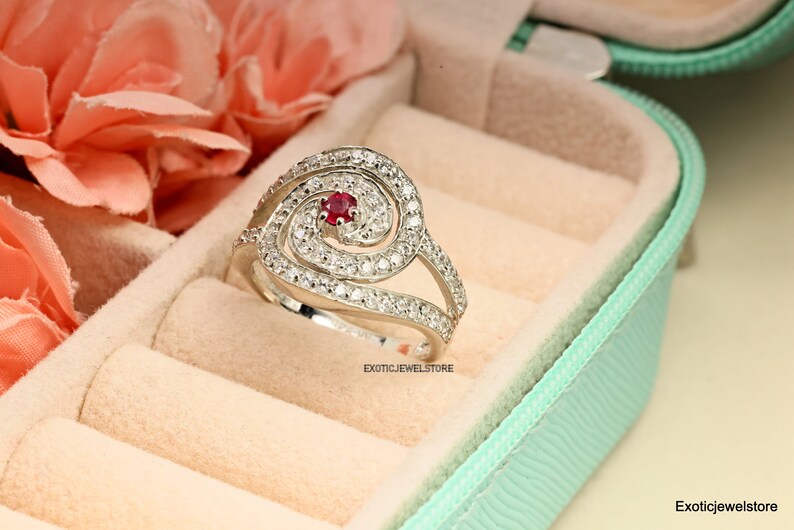 May include: A silver ring with a spiral design and a red gemstone in the center. The ring is set with small white gemstones.
