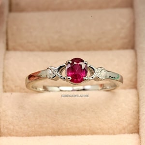 May include: A silver ring with a red oval gemstone and two smaller gemstones on either side. The ring is in a ring box.