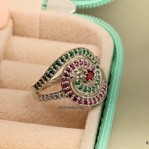 May include: A silver ring with a spiral design set with green, pink, and red gemstones. The ring is in a ring box.