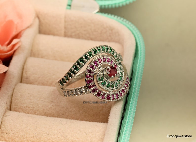May include: A silver ring with a spiral design set with green, pink, and red gemstones. The ring is in a ring box.