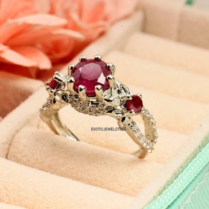 Op de afbeelding: Een zilveren ring met een grote, ronde, dieprode edelsteen als middelpunt, geflankeerd door twee kleinere rode stenen. De ring is bezet met kleinere, heldere stenen en wordt gepresenteerd in een lichtgroene sieradendoos.