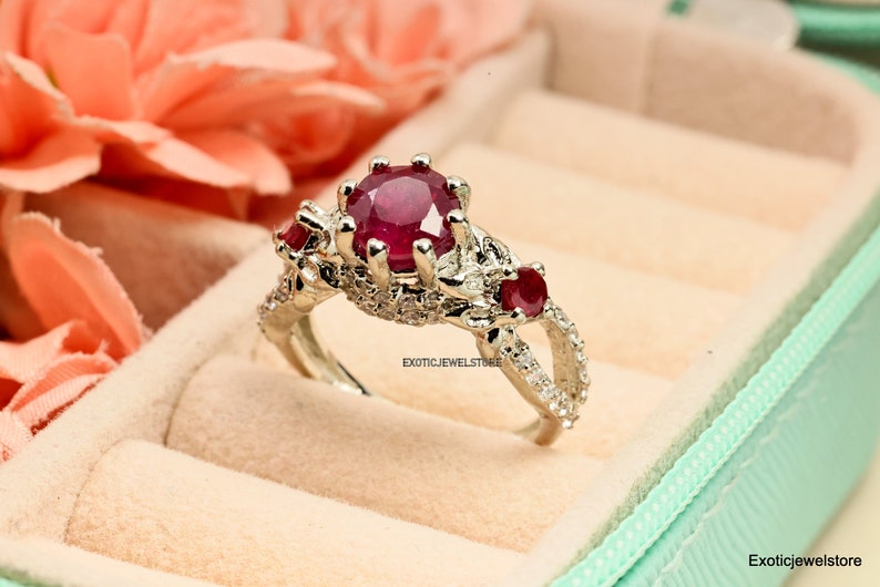 Op de afbeelding: Een zilveren ring met een grote, ronde, dieprode edelsteen als middelpunt, geflankeerd door twee kleinere rode stenen. De ring is bezet met kleinere, heldere stenen en wordt gepresenteerd in een lichtgroene sieradendoos.