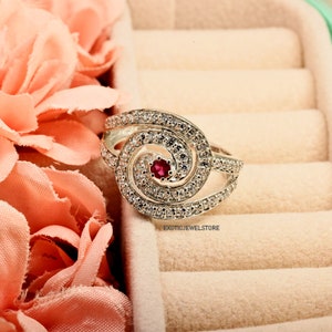 May include: A silver ring with a swirl design and a red gemstone in the center. The ring is in a light pink velvet ring box.