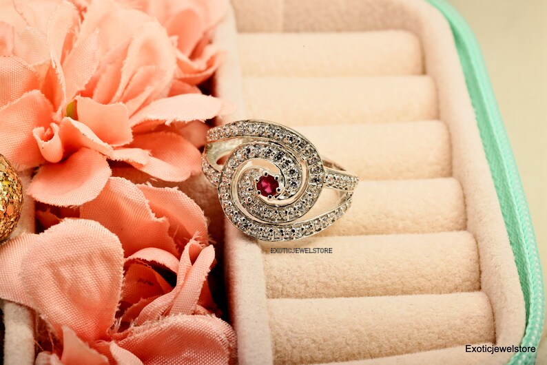 May include: A silver ring with a swirl design and a red gemstone in the center. The ring is in a light pink velvet ring box.