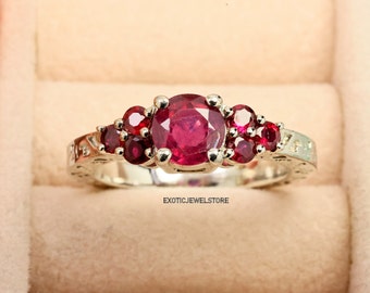 Puede incluir: Un anillo de plata con una gran piedra de rubí roja rodeada de piedras de rubí rojas más pequeñas. El anillo tiene una banda decorativa con un diseño floral.