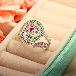 May include: A silver ring with a spiral design set with green and pink gemstones. The ring is in a light green ring box.