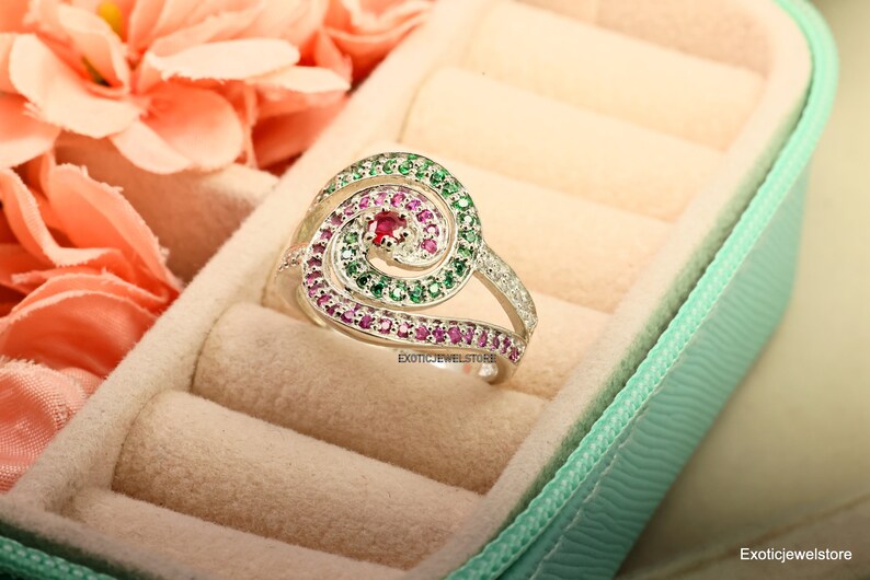 May include: A silver ring with a spiral design set with green and pink gemstones. The ring is in a light green ring box.