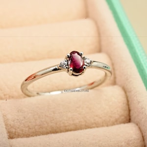 May include: A silver ring with a red oval gemstone and two smaller white gemstones on either side. The ring is in a light green ring box.