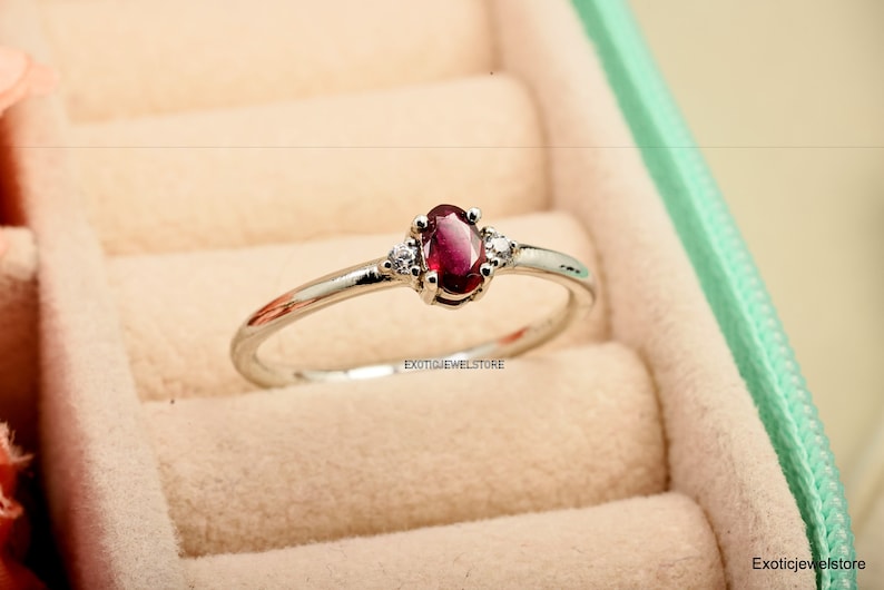 May include: A silver ring with a red oval gemstone and two smaller white gemstones on either side. The ring is in a light green ring box.