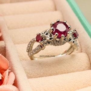 Op de afbeelding: Een zilveren ring met een grote, ronde, dieprode robijn als middensteen, geflankeerd door twee kleinere robijnen. De band heeft een gespleten ontwerp met kleine, heldere accentstenen. De ring wordt in een sieradendoos gepresenteerd.