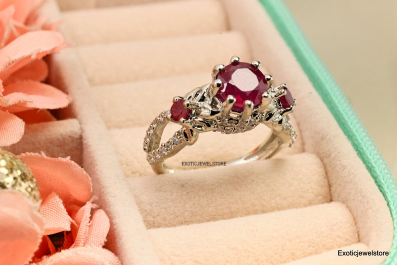 Op de afbeelding: Een zilveren ring met een grote, ronde, dieprode robijn als middensteen, geflankeerd door twee kleinere robijnen. De band heeft een gespleten ontwerp met kleine, heldere accentstenen. De ring wordt in een sieradendoos gepresenteerd.
