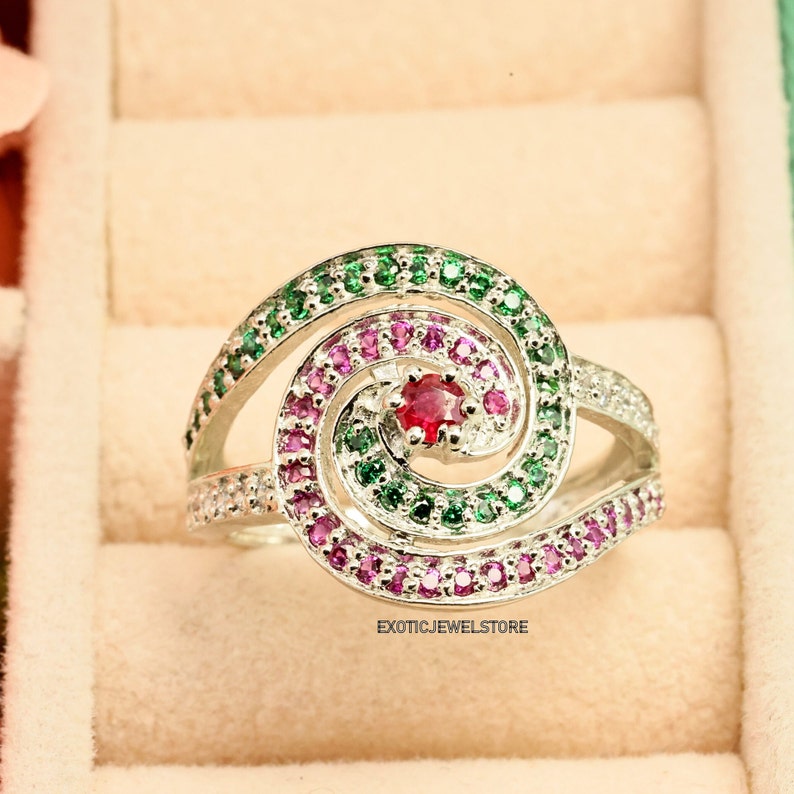 May include: A silver ring with a spiral design set with green and pink gemstones. A single red gemstone is at the center of the spiral.