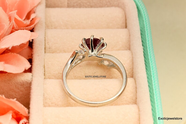 Op de afbeelding: Een zilveren ring met een grote rode edelsteen in het midden. De ring heeft een delicaat ontwerp met kleinere edelstenen op de band. De ring ligt in een ringendoos.