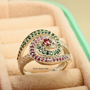 May include: A silver ring with a spiral design set with green and pink gemstones. The ring is in a jewelry box.