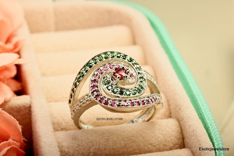 May include: A silver ring with a spiral design set with green and pink gemstones. The ring is in a jewelry box.