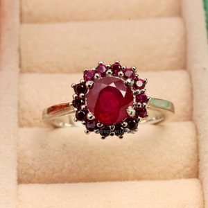 May include: A silver ring with a large, round, red ruby gemstone surrounded by smaller red rubies.