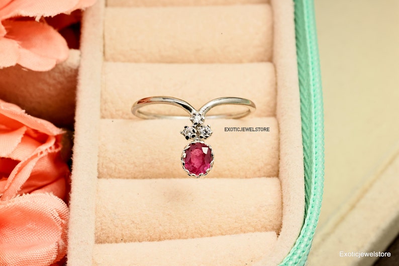 Pode incluir: Um anel de prata com uma gema de rubi rosa e tr&ecirc;s pequenos diamantes. O anel est&aacute; em uma caixa de anel verde claro com um interior branco.