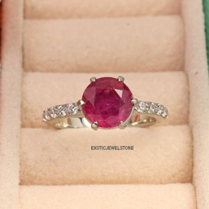Könnte beinhalten: Ein silberner Ring mit einem großen, runden, roten Rubin-Edelstein, der von einer Reihe kleiner, weißer Diamanten umgeben ist. Der Ring befindet sich in einer kleinen, weißen Schachtel mit dem Text "EXOTICJEWELSTORE" auf dem Boden.