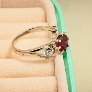 Op de afbeelding: Een zilveren ring met een grote rode edelsteen en twee kleinere witte edelstenen. De ring ligt in een fluwelen ringdoos.