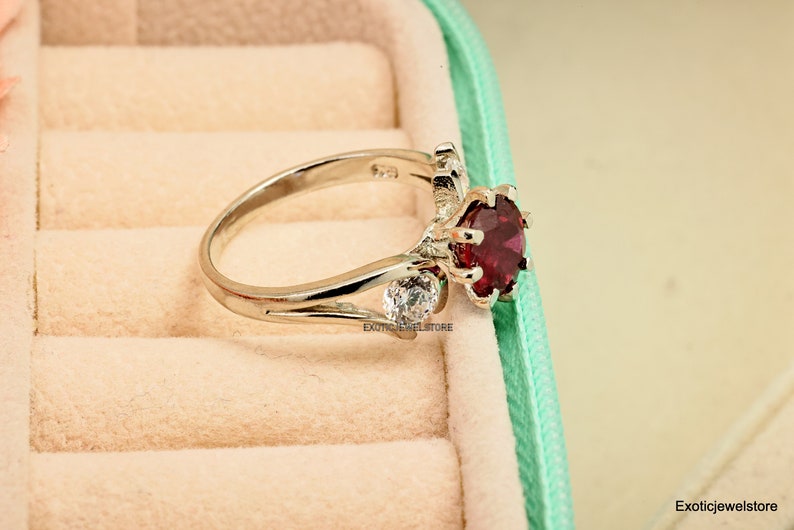 Op de afbeelding: Een zilveren ring met een grote rode edelsteen en twee kleinere witte edelstenen. De ring ligt in een fluwelen ringdoos.