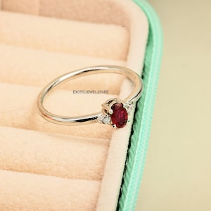May include: A silver ring with a red oval gemstone and smaller clear gemstones on the band. The ring is in a light green and cream colored ring box.