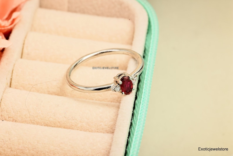 May include: A silver ring with a red oval gemstone and smaller clear gemstones on the band. The ring is in a light green and cream colored ring box.