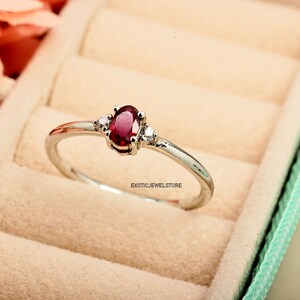 May include: A silver ring with a red oval gemstone and two smaller white gemstones. The ring is in a light green ring box.