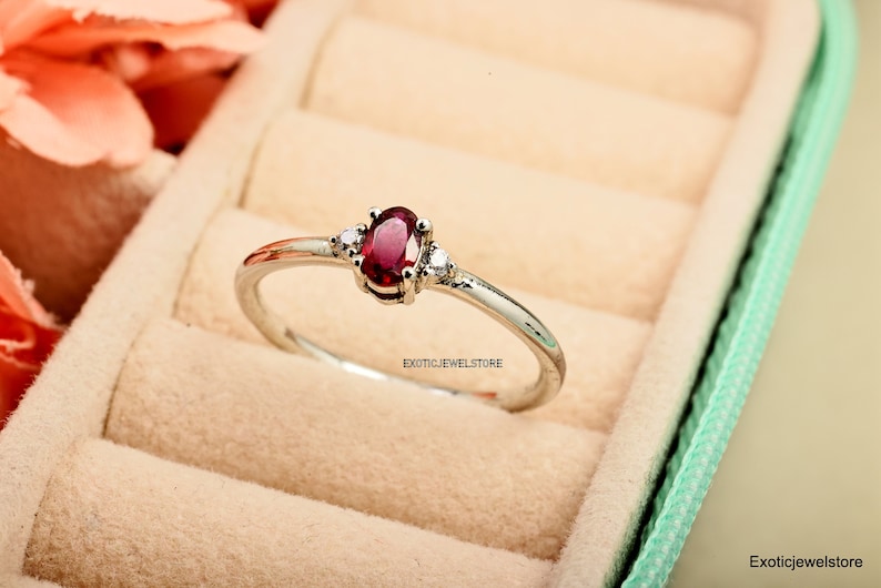 May include: A silver ring with a red oval gemstone and two smaller white gemstones. The ring is in a light green ring box.