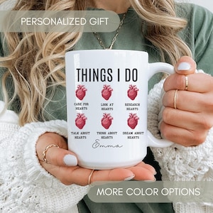 May include: White ceramic mug with a black and red design. The mug says "Things I Do" and has a heart graphic with the words "Care for Hearts", "Look at Hearts", "Research Hearts", "Talk About Hearts", "Think About Hearts", and "Dream About Hearts". The mug is personalized with the name "Emma".