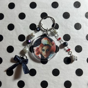 May include: A clear plastic keychain with a round photo insert featuring a person with red hair and a floral background. The keychain has a silver ring, white and black beads, a blue bow, and a silver dice charm.