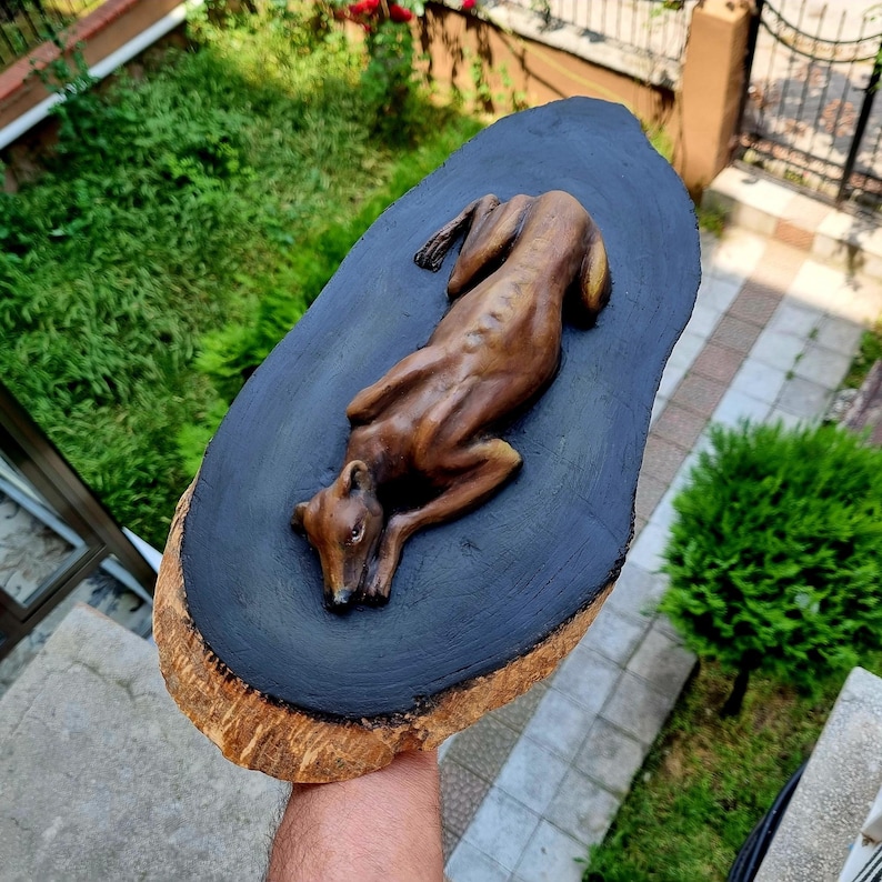 May include: A hand-carved wooden sculpture of a dog lying down, finished in a warm brown tone. The figure is mounted on a natural, oval-shaped wood slice with a dark black painted center and a textured, light brown bark edge surrounding it.