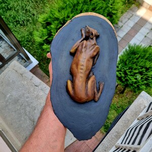 May include: A brown, sculpted figure of a dog in a resting position mounted on a dark, irregular, oval-shaped wooden plaque with a natural light-colored edge. The dog is depicted from a top-down perspective with its head turned toward its back.