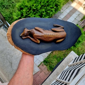 May include: A brown, sculpted figure of a greyhound dog lying down in a resting position, mounted onto a dark black, oval-shaped wooden plaque with a natural, light-colored bark edge visible along the side of the wood piece.