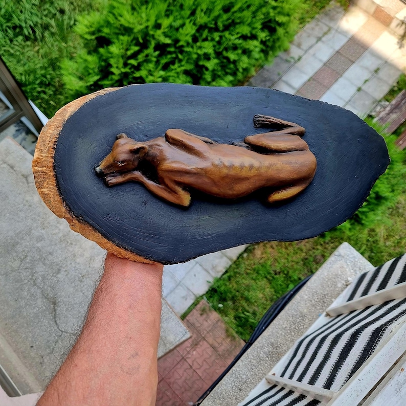 May include: A brown, sculpted figure of a greyhound dog lying down in a resting position, mounted onto a dark black, oval-shaped wooden plaque with a natural, light-colored bark edge visible along the side of the wood piece.