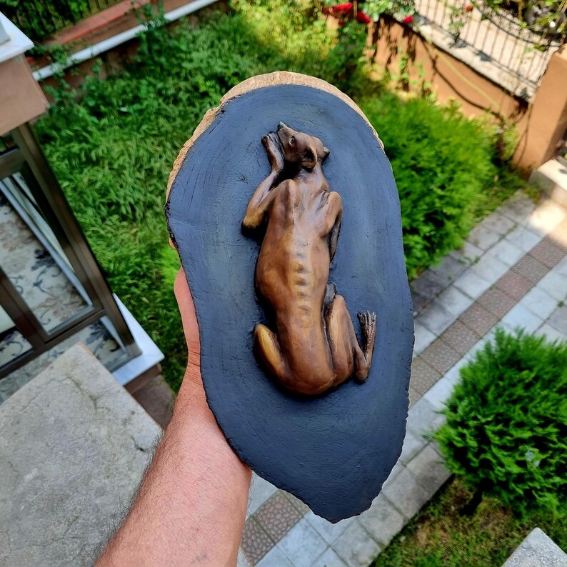May include: A brown, sculpted relief of a greyhound-like dog in a resting position, mounted on an oval-shaped, dark gray painted wooden slice with a natural light-colored wood edge. The dog figure is detailed with visible ribs and limbs.