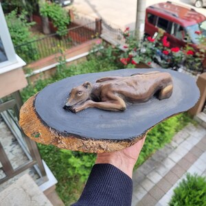 May include: A brown sculpted dog figure in a resting position, mounted on a flat, oval-shaped wooden slice. The top surface of the wood is painted a solid dark gray, while the outer edge retains the natural, textured brown tree bark.