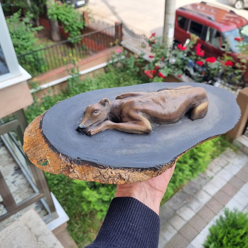 May include: A brown sculpted dog figure in a resting position, mounted on a flat, oval-shaped wooden slice. The top surface of the wood is painted a solid dark gray, while the outer edge retains the natural, textured brown tree bark.