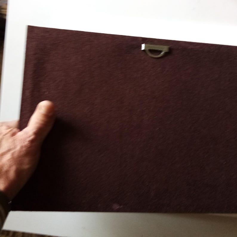 May include: A close-up of the back of a framed artwork. The frame backing is a dark maroon fabric. A metal D-ring is attached for hanging. A hand is holding the frame.