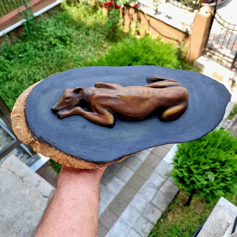 May include: A hand-carved wooden sculpture of a resting dog with a smooth brown finish, mounted on a flat, oval-shaped wooden slice with a dark black painted surface and natural bark edges, showcasing detailed anatomical features of the canine.