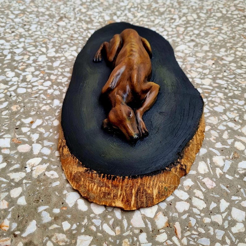 May include: A brown, sculpted dog figure lying down, mounted on a dark, circular wooden slice with visible bark edges. The dog is rendered in a realistic, three-dimensional style, contrasting against the smooth, dark-painted center of the wood.