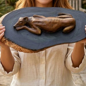 May include: A bronze-colored sculpture of a resting greyhound dog mounted on a dark, oval-shaped wooden plaque. The plaque features a smooth, black-painted center surface with natural, textured bark edges visible around the perimeter of the wood.