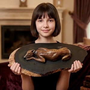 May include: A brown, sculpted figure of a dog in a resting position, mounted on a dark, oval-shaped wooden plaque with a natural, light-colored bark edge. The dog is depicted lying down with its head resting on its front paws.