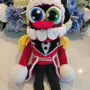 May include: A crocheted nutcracker doll featuring a red jacket with yellow fringe epaulets, black trousers, and a black hat. The doll has large mismatched blue and green eyes, a red nose, and a wide mouth with prominent white crocheted teeth.