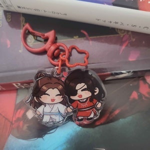 Può includere: Due personaggi in stile anime su un portachiavi rosso e bianco. I personaggi sono un ragazzo con i capelli neri e una ragazza con i capelli castani. Il ragazzo indossa un abito rosso e bianco e la ragazza indossa un abito bianco e blu. Il portachiavi è attaccato a una clip rossa e bianca.