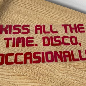May include: A red vinyl decal featuring the text KISS ALL THE TIME. DISCO, OCCASIONALLY in a blocky, pixelated font style. The letters are individual cutouts arranged in three lines, showcasing a bold, solid red colour throughout the design.