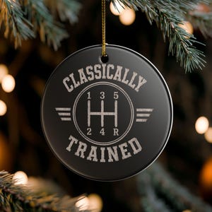 May include: A round black ceramic ornament featuring a white graphic of a manual transmission gear shift pattern with the numbers 1, 2, 3, 4, 5, and R. The text CLASSICALLY TRAINED is printed in a circular arc around the gear shift design.