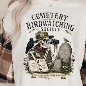 Könnte beinhalten: Ein weißes T-Shirt mit einer Grafik eines Skeletts mit Hut und Fernglas. Der Text lautet CEMETERY BIRDWATCHING SOCIETY, OBSERVED NOT DISTURBED, REST WELL FRIEND und Binoculars at the Burial Grounds, ergänzt durch Vögel und Grabsteine.