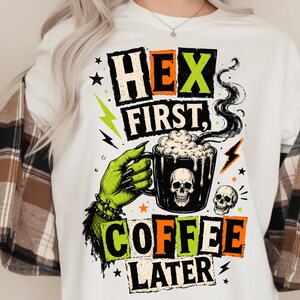 Può includere: Una t-shirt bianca con una stampa grafica di una mano verde con lunghe unghie nere che tiene una tazza nera con un teschio e schiuma fumante. Il testo recita HEX FIRST, COFFEE LATER in lettere stampatello arancioni e verdi.